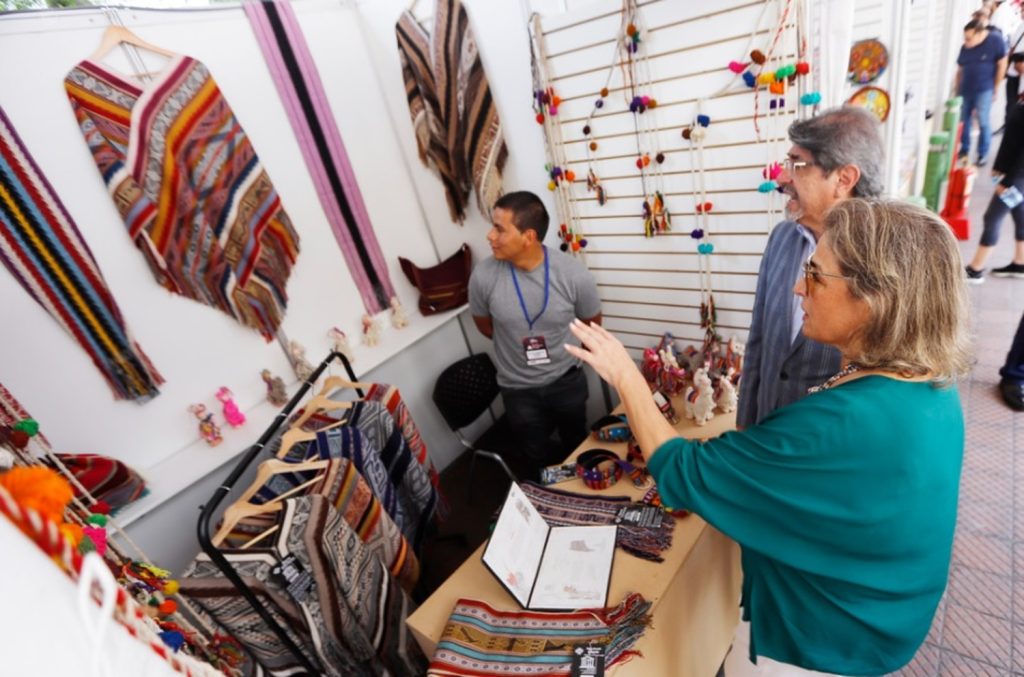 Visitantes recorren un stand de artesanías textiles durante una feria, observando y apreciando ponchos, chalinas, tapices y accesorios elaborados con técnicas tradicionales. La imagen refleja el valor del trabajo artesanal, el intercambio entre productores y compradores, y la promoción del patrimonio cultural a través de piezas que destacan por sus colores, diseños y acabados hechos a mano.